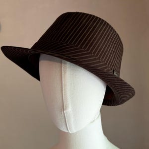 K&ouml;nnte beinhalten: Ein dunkelbrauner Fedora-Hut mit einem dezenten vertikalen Nadelstreifenmuster auf der Krone und der Krempe. Der Hut wird auf einem schlichten wei&szlig;en Schaufensterpuppenkopf pr&auml;sentiert und zeigt die strukturierte Form und das Stoffdesign.