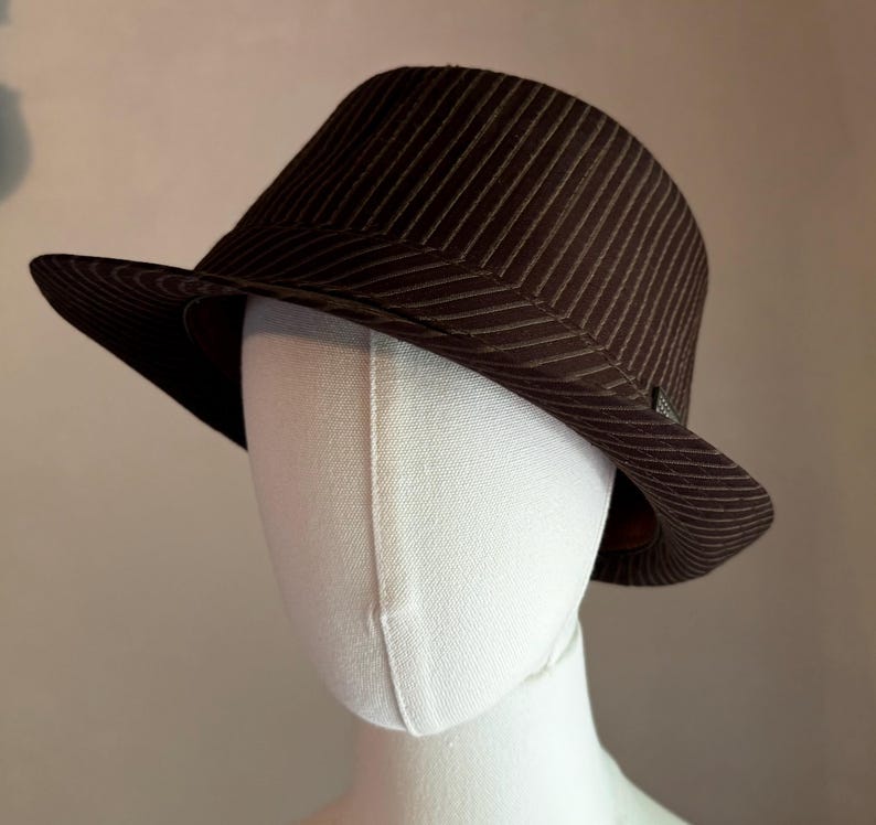 K&ouml;nnte beinhalten: Ein dunkelbrauner Fedora-Hut mit einem dezenten vertikalen Nadelstreifenmuster auf der Krone und der Krempe. Der Hut wird auf einem schlichten wei&szlig;en Schaufensterpuppenkopf pr&auml;sentiert und zeigt die strukturierte Form und das Stoffdesign.