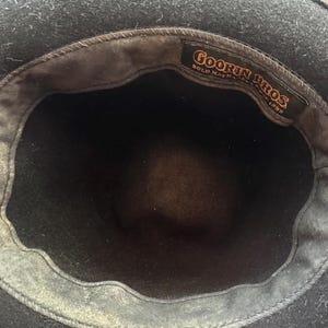May include: A black hat with a dark interior lining. A rectangular fabric label is stitched inside the crown, featuring the text Goorin Bros. Bold Hatters Since 1895 in orange lettering against a black background. The hat has a wide brim.