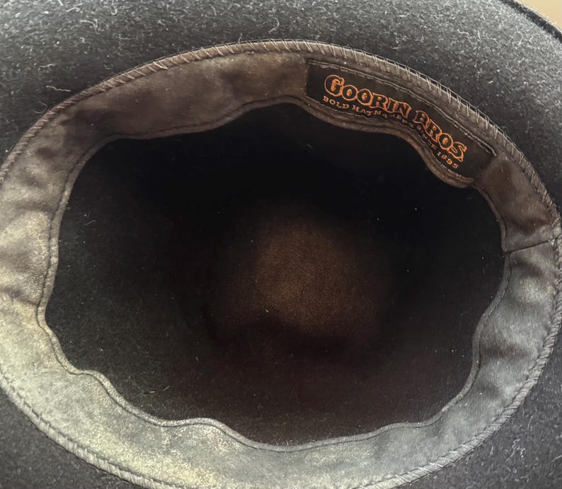 May include: A black hat with a dark interior lining. A rectangular fabric label is stitched inside the crown, featuring the text Goorin Bros. Bold Hatters Since 1895 in orange lettering against a black background. The hat has a wide brim.