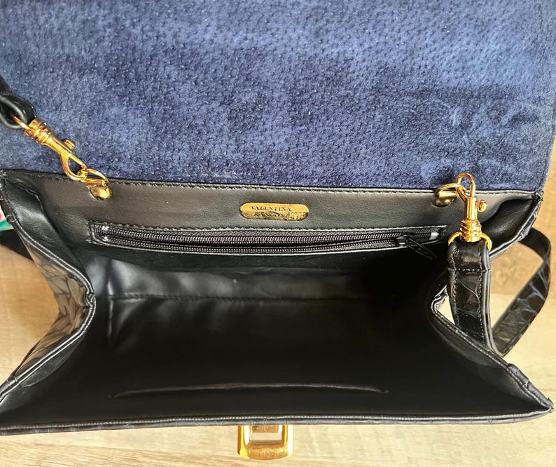 May include: Black leather handbag with a gold chain strap and a gold clasp. The bag has a blue suede lining and a label that reads "Valentino."