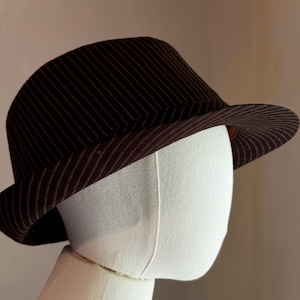 K&ouml;nnte beinhalten: Ein dunkelbrauner Fedora-Hut mit einem dezenten Nadelstreifenmuster auf der Krone und der Krempe. Der Hut wird auf einem schlichten wei&szlig;en Schaufensterpuppenkopf pr&auml;sentiert und zeigt die strukturierte Form und die nach unten geneigte Krempe.