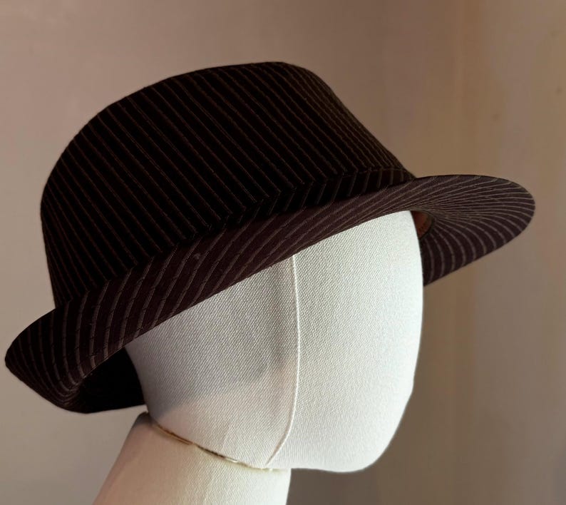 K&ouml;nnte beinhalten: Ein dunkelbrauner Fedora-Hut mit einem dezenten Nadelstreifenmuster auf der Krone und der Krempe. Der Hut wird auf einem schlichten wei&szlig;en Schaufensterpuppenkopf pr&auml;sentiert und zeigt die strukturierte Form und die nach unten geneigte Krempe.