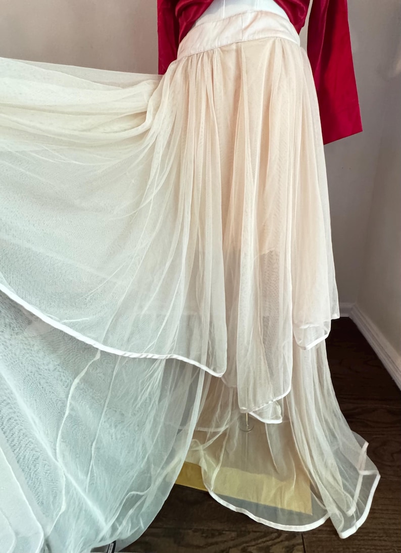K&ouml;nnte beinhalten: Ein mehrlagiges, flie&szlig;endes Kleid mit einem cremefarbenen T&uuml;llrock und einem roten Samtoberteil. Der Rock hat mehrere Lagen aus transparentem Stoff, wodurch ein volumin&ouml;ser Effekt entsteht. Das Kleid ist f&uuml;r einen besonderen Anlass.