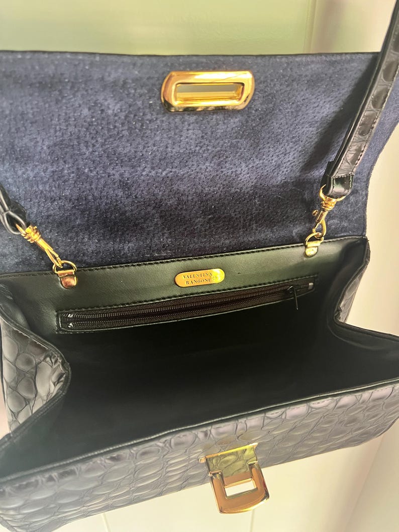 May include: Black crocodile embossed leather handbag with a gold clasp and a gold zipper. The bag has a black fabric lining and a gold label that reads "VALENTINO  B.ANGIONE".