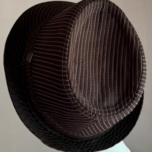 K&ouml;nnte beinhalten: Ein dunkelbrauner Fedora-Hut mit vertikaler Kordstruktur auf der Krone und einem kontrastierenden schwarzen Netzband mit Rautenmuster um die Basis. Der Hut hat eine strukturierte Form mit einer eingedr&uuml;ckten Oberseite und einem kleinen Detail.