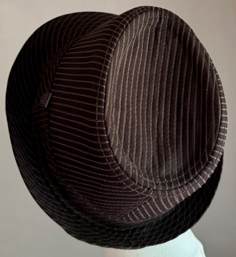 K&ouml;nnte beinhalten: Ein dunkelbrauner Fedora-Hut mit vertikaler Kordstruktur auf der Krone und einem kontrastierenden schwarzen Netzband mit Rautenmuster um die Basis. Der Hut hat eine strukturierte Form mit einer eingedr&uuml;ckten Oberseite und einem kleinen Detail.