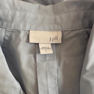 May include: Close-up of a light blue shirt collar with a label that reads "J.Jill" and "MADE IN PHILIPPINES L". The shirt is made of a soft, lightweight fabric.