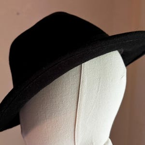 May include: A solid black fedora hat with a structured crown and a wide, slightly curved brim. The hat is displayed on a plain, off-white mannequin head form, highlighting the smooth texture of the material and the clean, simple silhouette.