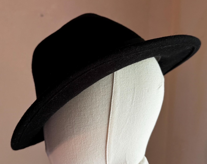 May include: A solid black fedora hat with a structured crown and a wide, slightly curved brim. The hat is displayed on a plain, off-white mannequin head form, highlighting the smooth texture of the material and the clean, simple silhouette.