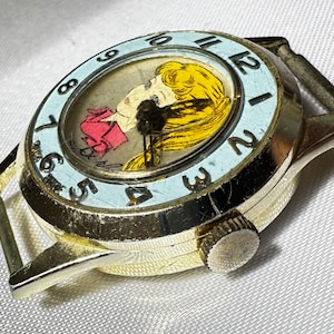 May include: A gold-tone wristwatch featuring a light blue bezel with black numerals. The watch face displays a graphic illustration of a person with yellow hair and a pink garment. The watch case is round with a side crown and metal lugs.