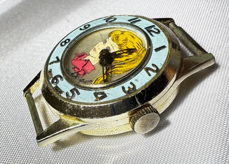 May include: A gold-tone wristwatch featuring a light blue bezel with black numerals. The watch face displays a graphic illustration of a person with yellow hair and a pink garment. The watch case is round with a side crown and metal lugs.