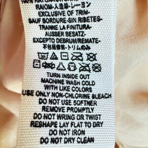 K&ouml;nnte beinhalten: Ein wei&szlig;es Stoffetikett mit schwarzem Text, das Pflegehinweise und Details zur Stoffzusammensetzung enth&auml;lt. Das Etikett enth&auml;lt Waschsymbole und S&auml;tze wie "Machine Wash Cold" und "Do Not Iron". Das Etikett befindet sich auf einem hellpfirsichfarbenen Stoff.