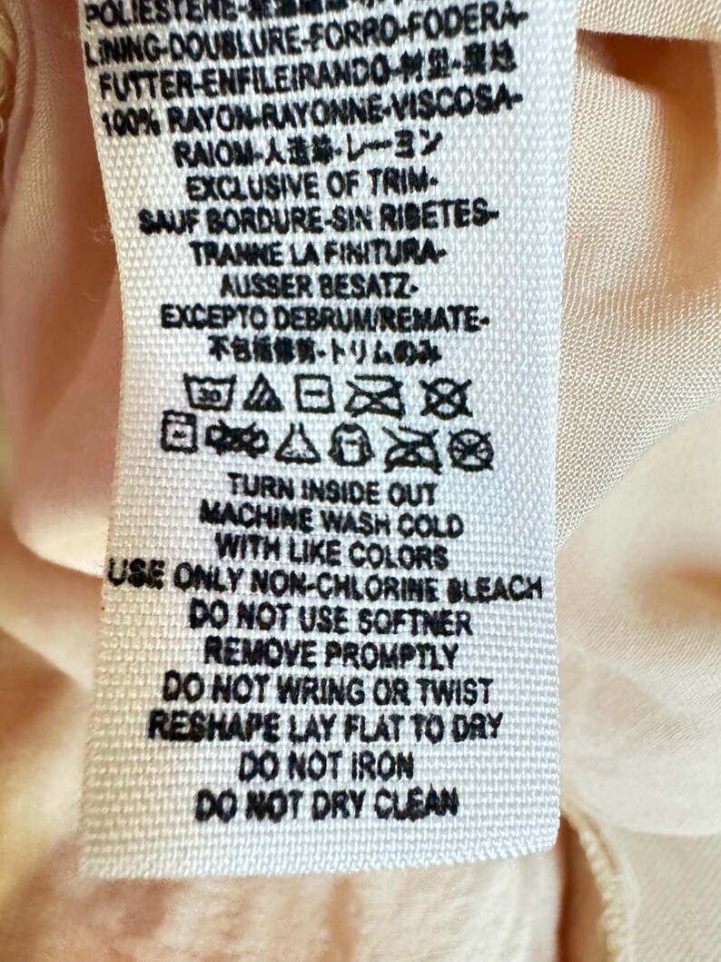 K&ouml;nnte beinhalten: Ein wei&szlig;es Stoffetikett mit schwarzem Text, das Pflegehinweise und Details zur Stoffzusammensetzung enth&auml;lt. Das Etikett enth&auml;lt Waschsymbole und S&auml;tze wie "Machine Wash Cold" und "Do Not Iron". Das Etikett befindet sich auf einem hellpfirsichfarbenen Stoff.