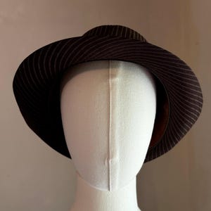 K&ouml;nnte beinhalten: Ein dunkelbrauner Fedora-Hut mit einem dezenten, d&uuml;nnen, hellen Nadelstreifenmuster auf der Krone und der breiten, leicht nach unten geneigten Krempe. Der Hut wird auf einem schlichten, cremewei&szlig;en, gesichtslosen Schaufensterpuppenkopf pr&auml;sentiert.