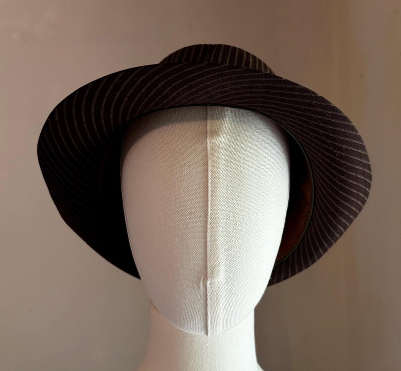 K&ouml;nnte beinhalten: Ein dunkelbrauner Fedora-Hut mit einem dezenten, d&uuml;nnen, hellen Nadelstreifenmuster auf der Krone und der breiten, leicht nach unten geneigten Krempe. Der Hut wird auf einem schlichten, cremewei&szlig;en, gesichtslosen Schaufensterpuppenkopf pr&auml;sentiert.