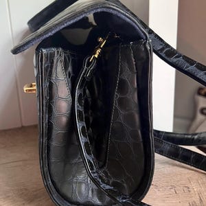May include: Black leather handbag with a crocodile embossed pattern and a gold-tone clasp. The bag has a long strap and a top handle.
