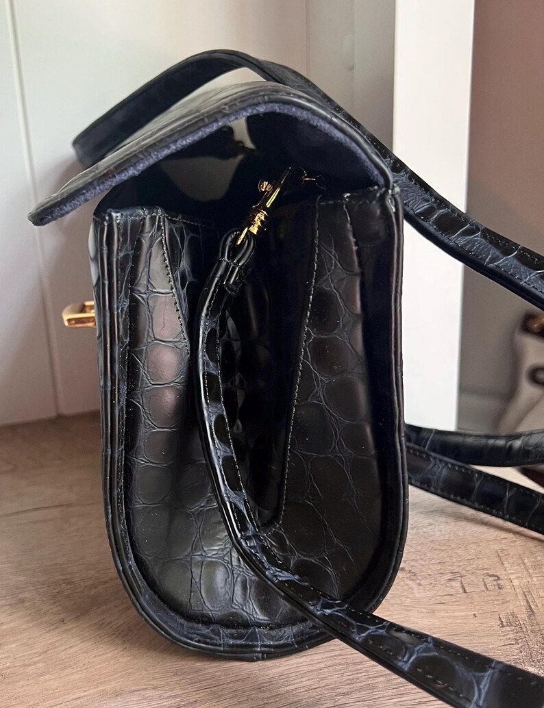 May include: Black leather handbag with a crocodile embossed pattern and a gold-tone clasp. The bag has a long strap and a top handle.