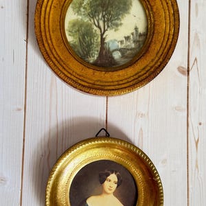 May include: Two antique framed miniature paintings. The top painting depicts a landscape with a tree, bridge, and buildings. The bottom painting is a portrait of a woman in a blue dress. Both paintings are in round, gold-colored frames.