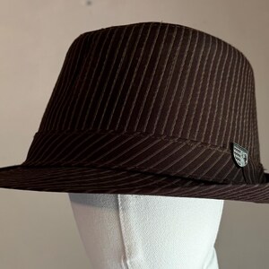 K&ouml;nnte beinhalten: Ein dunkelbrauner Fedora-Hut mit einem dezenten vertikalen Nadelstreifenmuster auf der Krone und der Krempe. Der Hut verf&uuml;gt &uuml;ber ein passendes braunes Band um die Basis und ein kleines, silberfarbenes Metallemblem an der Seite des Bandes.
