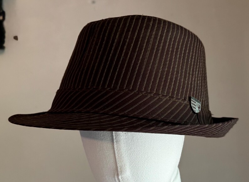 K&ouml;nnte beinhalten: Ein dunkelbrauner Fedora-Hut mit einem dezenten vertikalen Nadelstreifenmuster auf der Krone und der Krempe. Der Hut verf&uuml;gt &uuml;ber ein passendes braunes Band um die Basis und ein kleines, silberfarbenes Metallemblem an der Seite des Bandes.