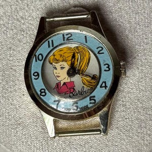 May include: A gold-tone analog wristwatch featuring a light blue dial with black numerals from 1 to 12. The center of the dial displays a graphic illustration of a blonde character with a ponytail wearing a pink shirt, with the Barbie logo printed below.