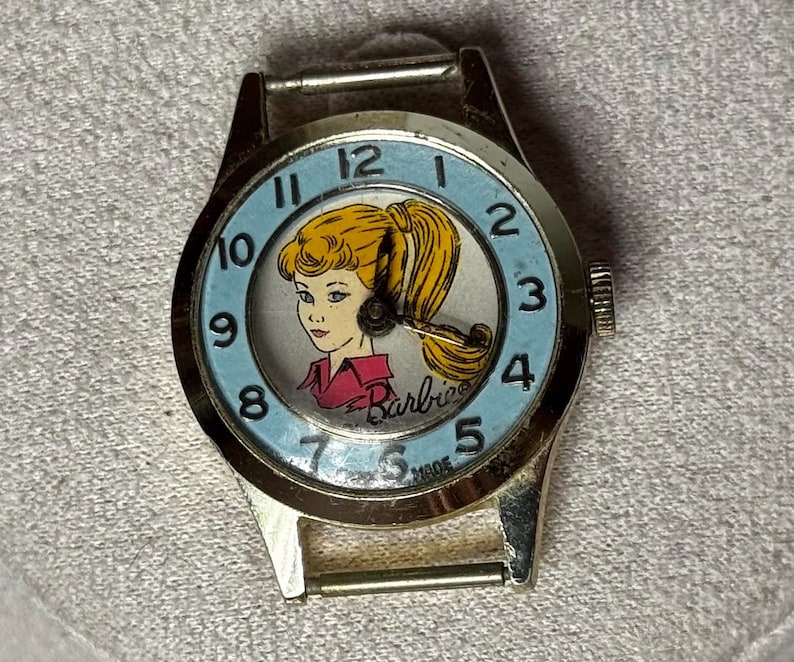 May include: A gold-tone analog wristwatch featuring a light blue dial with black numerals from 1 to 12. The center of the dial displays a graphic illustration of a blonde character with a ponytail wearing a pink shirt, with the Barbie logo printed below.