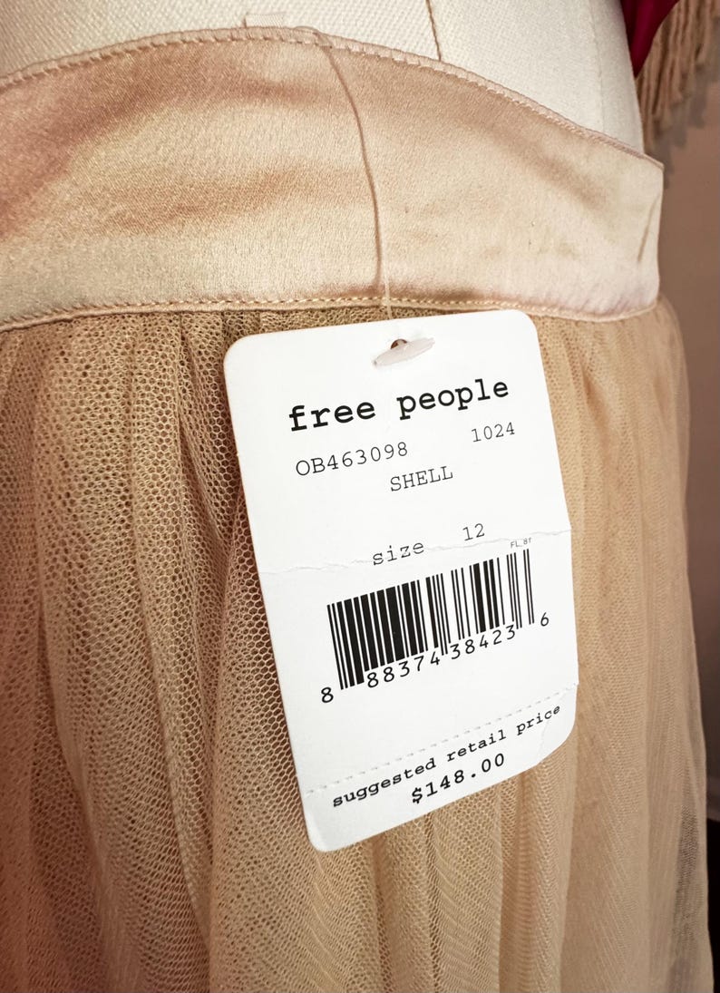 May include: A close-up of a beige skirt with a tulle overlay. The skirt has a satin waistband and a tag that reads "free people", size 12, and "SHELL". The suggested retail price is $148.00.