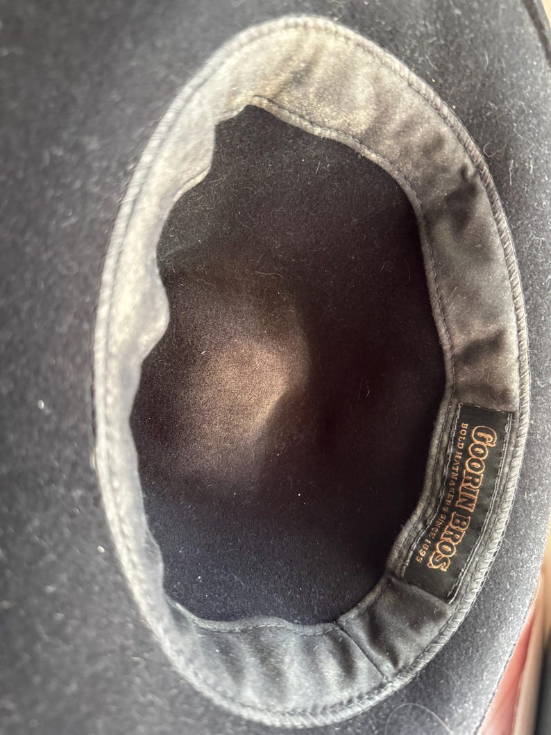 May include: A black hat viewed from the interior, featuring a dark fabric lining and a rectangular black label stitched into the inner band. The label displays the gold text Goorin Bros. Bold Hatmakers Since 1895 in a decorative serif font.