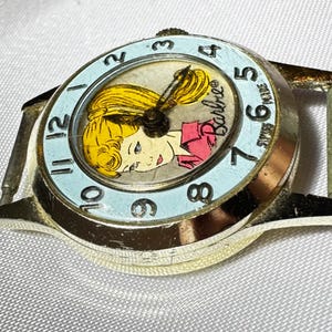 May include: A gold-tone analog wristwatch featuring a light blue bezel with black numerals from 1 to 12. The watch face displays a graphic illustration of a blonde character with the text Barbie and Swiss Made printed in black on the dial.