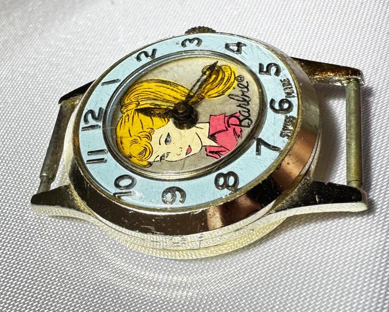 May include: A gold-tone analog wristwatch featuring a light blue bezel with black numerals from 1 to 12. The watch face displays a graphic illustration of a blonde character with the text Barbie and Swiss Made printed in black on the dial.