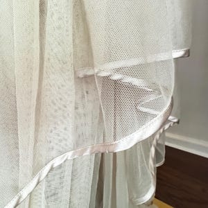 K&ouml;nnte beinhalten: Nahaufnahme eines mehrschichtigen, cremefarbenen T&uuml;llstoffs mit zarter, netzartiger Textur. Die Kanten sind mit einem Satinband in einer passenden hellen Farbe verziert, wodurch ein weicher, flie&szlig;ender Effekt entsteht. Der Stoff scheint Teil eines Kleidungsst&uuml;cks zu sein.