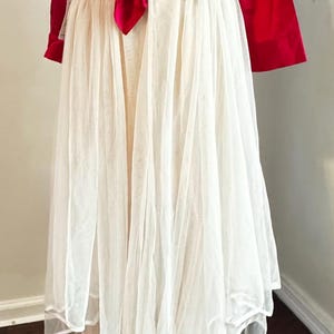 K&ouml;nnte beinhalten: Ein rotes Satin-Oberteil mit langen &Auml;rmeln und gebundener Front, kombiniert mit einem mehrschichtigen wei&szlig;en T&uuml;llrock. Der Rock hat ein flie&szlig;endes, mehrschichtiges Design. Das Outfit ist f&uuml;r besondere Anl&auml;sse geeignet.