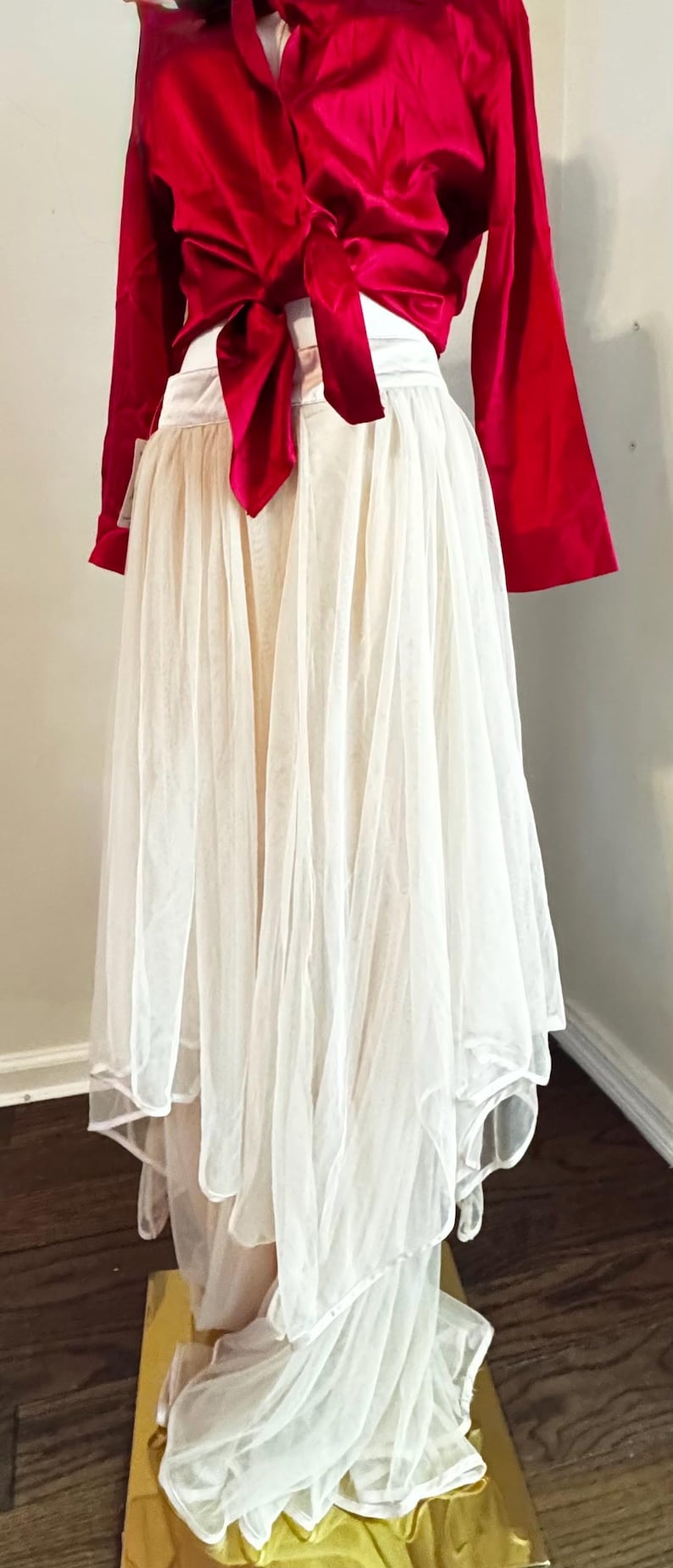 K&ouml;nnte beinhalten: Ein rotes Satin-Oberteil mit langen &Auml;rmeln und gebundener Front, kombiniert mit einem mehrschichtigen wei&szlig;en T&uuml;llrock. Der Rock hat ein flie&szlig;endes, mehrschichtiges Design. Das Outfit ist f&uuml;r besondere Anl&auml;sse geeignet.