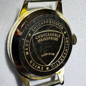 May include: A gold-tone watch case back with a black circular center featuring gold text that reads SHOCKPROTECTED, UNBREAKABLE MAINSPRING, ALUMINIUM, ANTIMAGNETIC, and SWISS MADE. The metal surface shows visible scratches and wear.