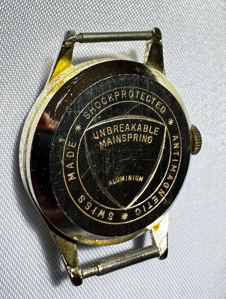 May include: A gold-tone watch case back with a black circular center featuring gold text that reads SHOCKPROTECTED, UNBREAKABLE MAINSPRING, ALUMINIUM, ANTIMAGNETIC, and SWISS MADE. The metal surface shows visible scratches and wear.