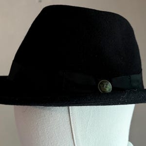 May include: A black fedora hat with a structured crown and a wide brim. The hat features a black grosgrain ribbon band encircling the base of the crown, accented with a small, circular metallic emblem on the side displaying a crown motif.