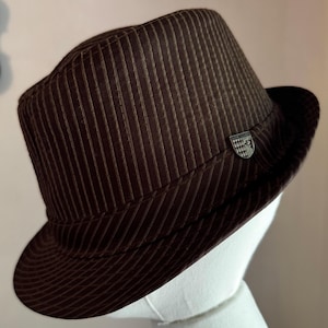 K&ouml;nnte beinhalten: Ein dunkelbrauner Fedora-Hut mit einem dezenten vertikalen Nadelstreifenmuster auf der Krone und der Krempe. Der Hut verf&uuml;gt &uuml;ber ein passendes braunes Stoffband um die Basis und ein kleines, silberfarbenes, schildf&ouml;rmiges Metallabzeichen an der Seite.