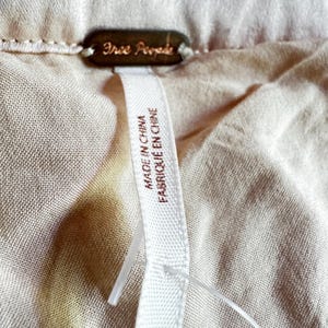 K&ouml;nnte beinhalten: Nahaufnahme eines beigefarbenen Stoffes mit einem Free People Markenetikett. Das Etikett hat eine dunkelbronze Farbe. Ein wei&szlig;es Etikett am Kleidungsst&uuml;ck tr&auml;gt die Aufschrift "MADE IN CHINA FABRIQU&Eacute; EN CHINE".
