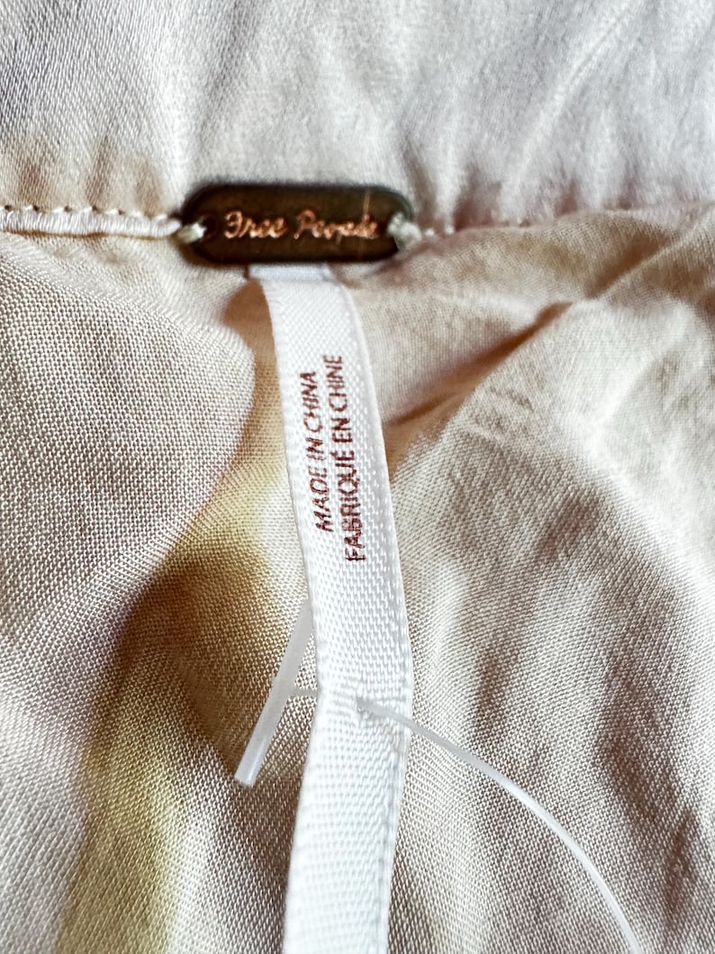 K&ouml;nnte beinhalten: Nahaufnahme eines beigefarbenen Stoffes mit einem Free People Markenetikett. Das Etikett hat eine dunkelbronze Farbe. Ein wei&szlig;es Etikett am Kleidungsst&uuml;ck tr&auml;gt die Aufschrift "MADE IN CHINA FABRIQU&Eacute; EN CHINE".