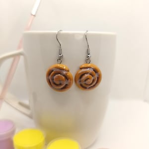 May include: A pair of dangle earrings featuring miniature cinnamon roll designs. The earrings are a warm brown color with a spiral pattern, suspended from silver-colored hooks. The earrings are set against a white background.