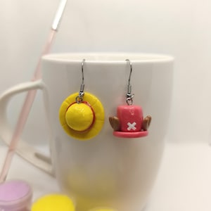 May include: A pair of handmade earrings featuring a yellow straw hat and a pink hat with a white cross. The earrings are suspended from silver-colored fish hook ear wires. The earrings are displayed in front of a white mug.
