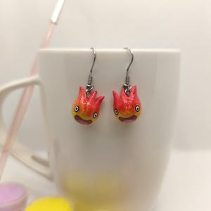 May include: A pair of handmade earrings featuring a cartoon fire demon design. The earrings are red and yellow with black eyes and a smiling mouth. They hang from silver-colored fish hook ear wires. The earrings are displayed in front of a white mug.