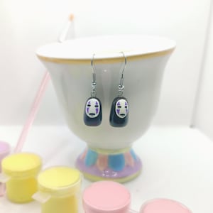 May include: A pair of silver dangle earrings featuring a black and white No Face character from the animated film Spirited Away. The character has purple accents on the face.