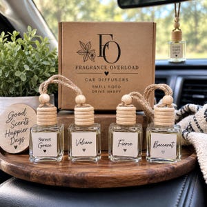 May include: Four square glass car diffuser bottles with wooden caps and jute rope hangers, labeled Sweet Grace, Volcano, Fierce, and Baccarat. They sit before a brown cardboard box printed with Fragrance Overload Car Diffusers Smell Good Drive Happy.