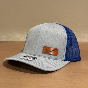 May include: A gray and blue trucker hat with a brown leather patch featuring a crane silhouette. The patch has the text "Crane Operator" on it.