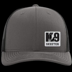 May include: A gray trucker hat with a black mesh back and a curved brim. The front features a white square patch with the text "K9 SKEETER" and a silhouette of a person and dog.
