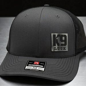 May include: Charcoal gray trucker hat with a black mesh back and a curved brim. The hat features a metal patch with the text "K9 LUUK". The Richardson 112 adjustable hat has a structured front.