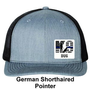 May include: A blue and black trucker hat with a graphic of a dog and the text "K9 DUG" on the front. The hat has a mesh back and a curved brim. The text "German Shorthaired Pointer" is below the hat.