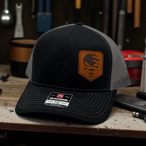 May include: Black and gray trucker hat with a leather patch featuring an American flag design. The hat has a curved brim and a mesh back. The patch is brown and the hat is adjustable.